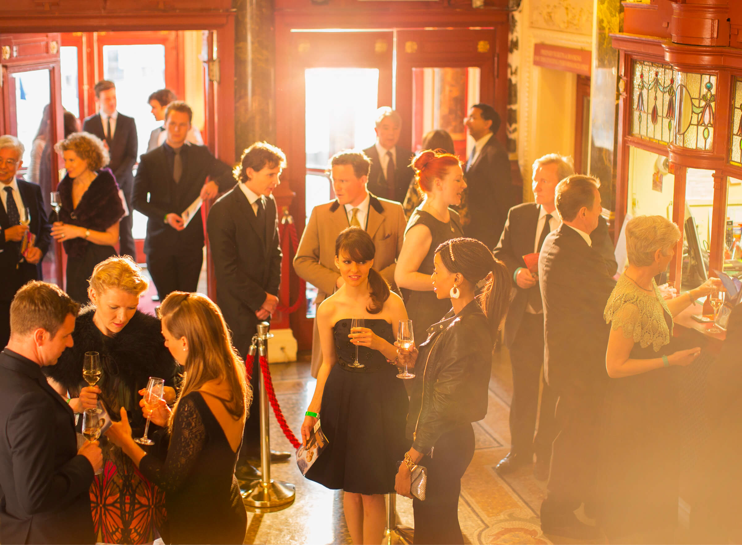 Theatergoers in evening dress mingling over drinks in a grand theater lobby